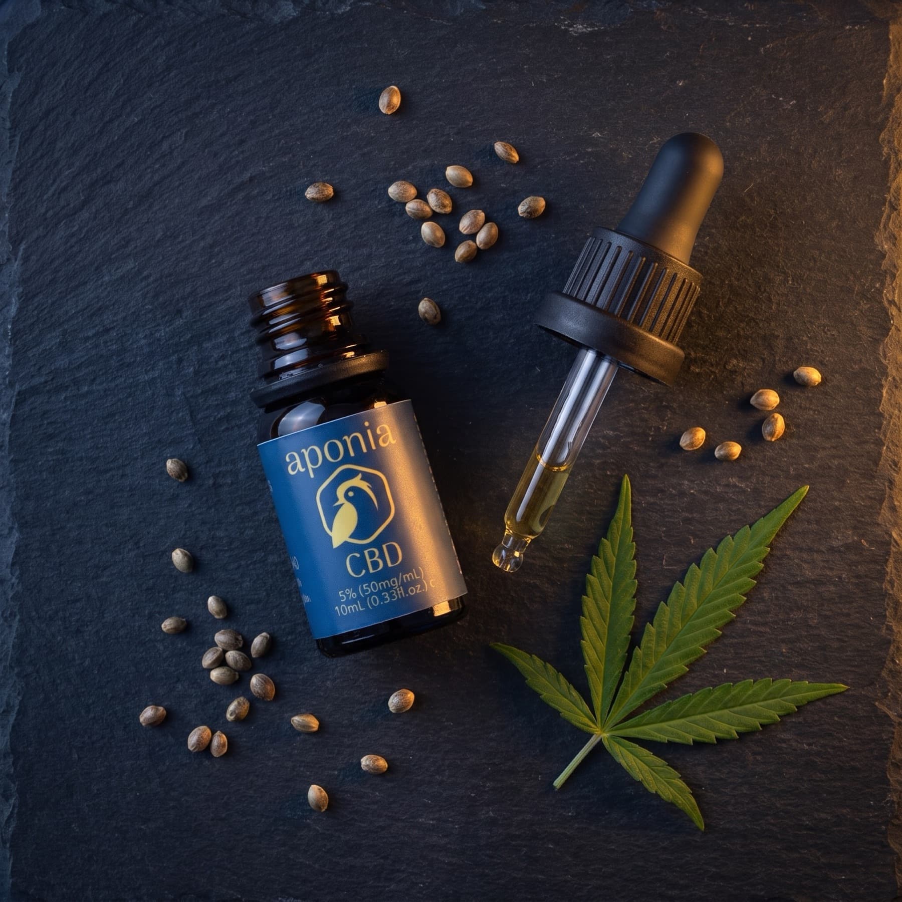 Aponia CBD oil bottle, dropper and scattered hemp seeds arranged on dark slate